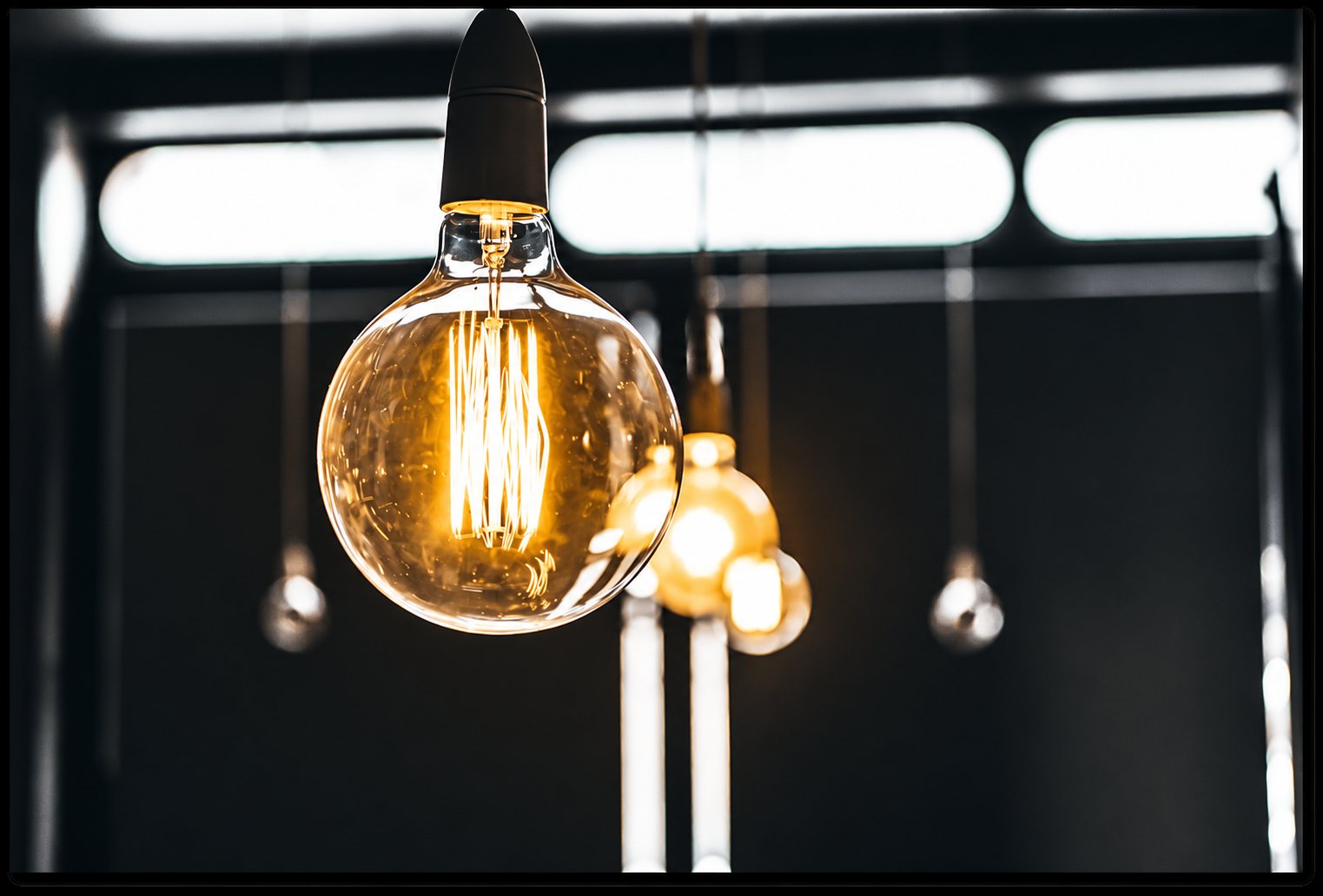 Une grosse ampoule globe à filament lumineux orangé est suspendue au premier plan, avec d’autres ampoules floues en arrière-plan dans un intérieur sombre.