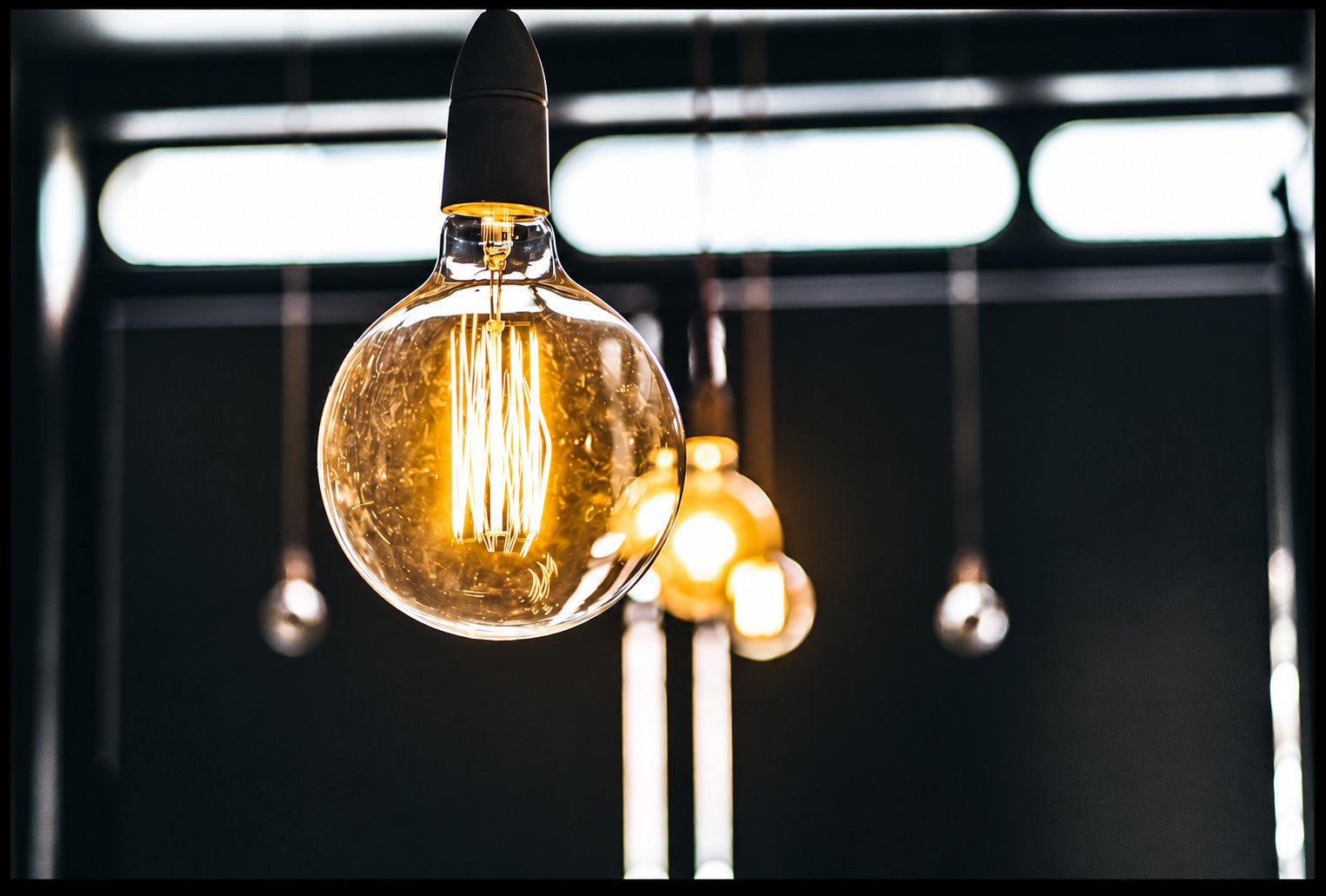 Gros plan sur une ampoule globe à filament incandescent suspendue, avec d’autres ampoules floues en arrière-plan dans un intérieur sombre.