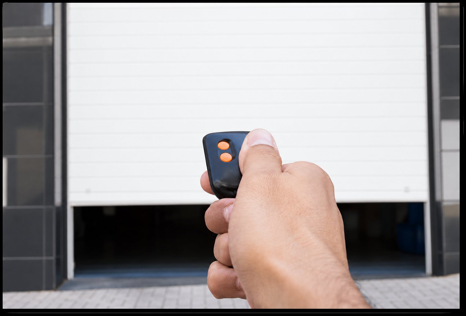 Une main tient une petite télécommande noire à deux boutons orange pour ouvrir une porte de garage blanche qui se lève.